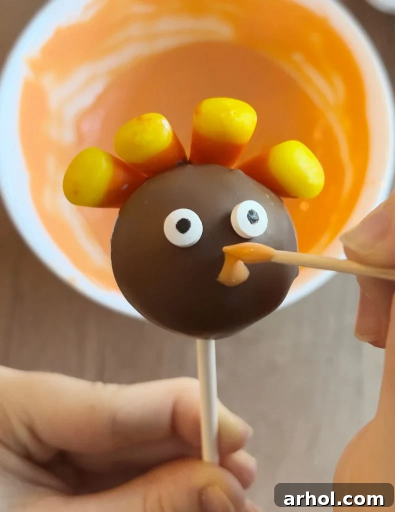 A toothpick is used to draw a small orange beak onto a chocolate-covered turkey cake pop's face.