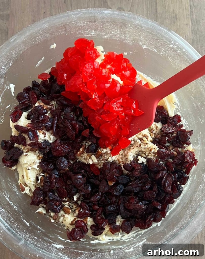Hands stirring chopped cherries and pecans into cookie dough with a spatula in a mixing bowl.