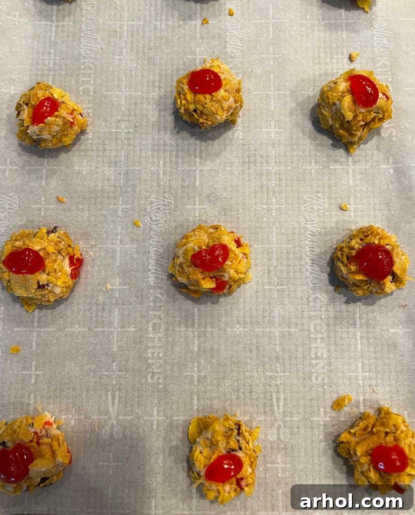 Unbaked cherry wink cookies, perfectly rolled in crushed cornflakes and topped with quartered cherries, arranged on parchment paper-lined baking sheets.