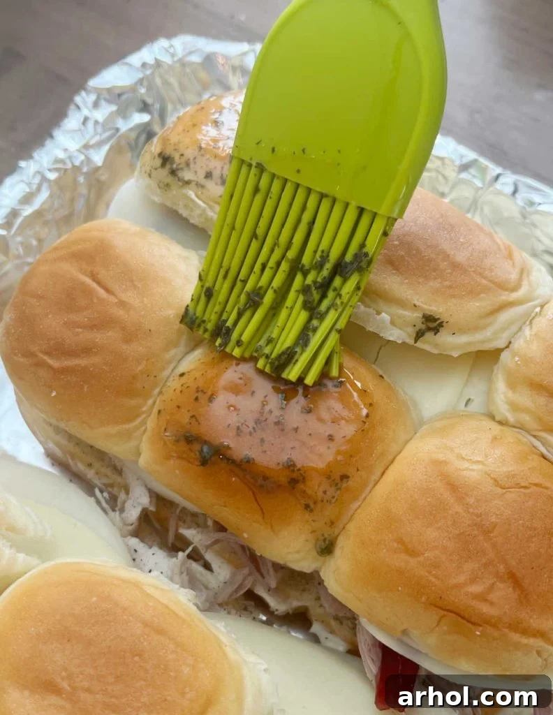 Aloha Turkey Sliders 6 Melted butter mixture being brushed onto the tops of assembled turkey cranberry sliders on Hawaiian rolls before baking.