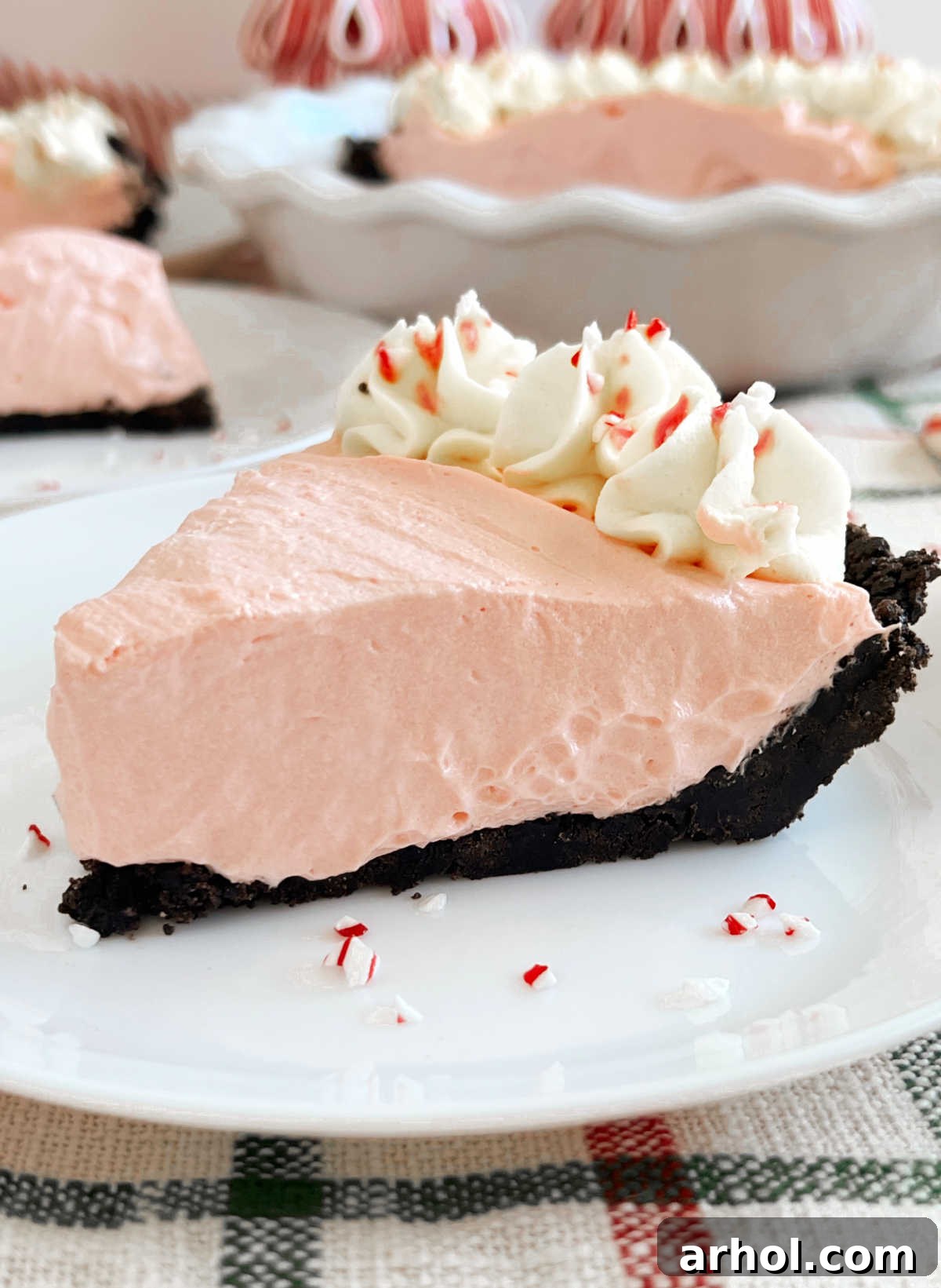 A slice of vibrant pink no-bake peppermint pie with an Oreo crust, garnished with whipped cream and crushed candy canes, on a white plate, ready to be enjoyed.