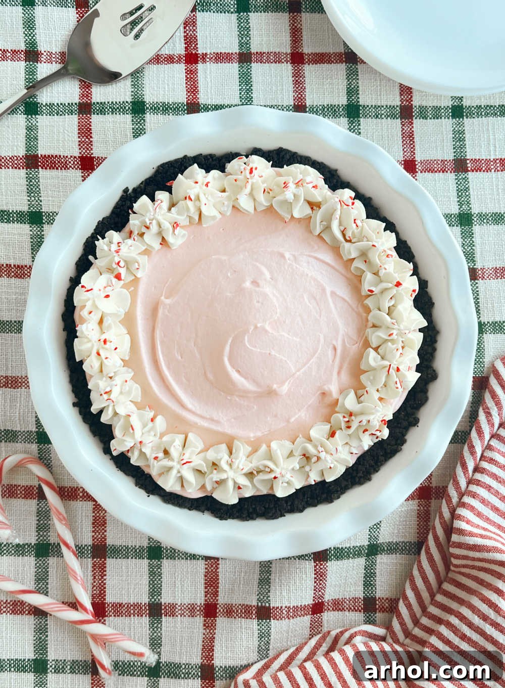 A whole peppermint pie with a luscious pink filling, topped with whipped cream stars and vibrant crushed peppermint pieces, sitting on a festive table.