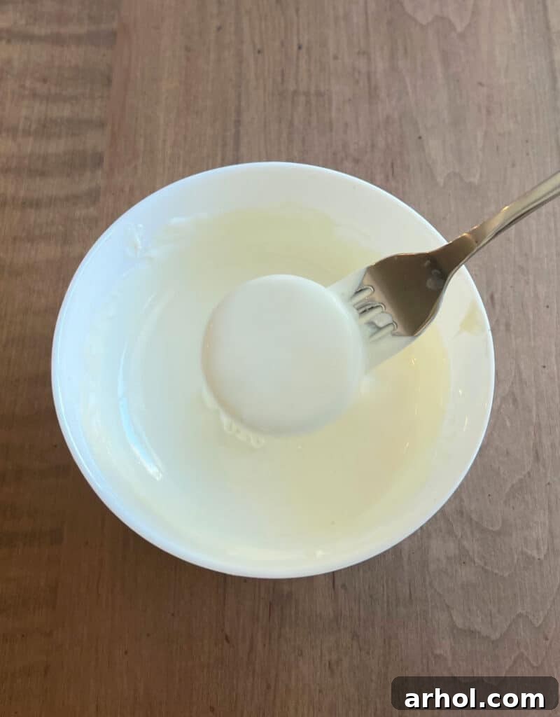 Irresistible White Chocolate Covered Oreos 4 Carefully dipping an Oreo cookie into a bowl of smooth white chocolate candy coating using a fork.