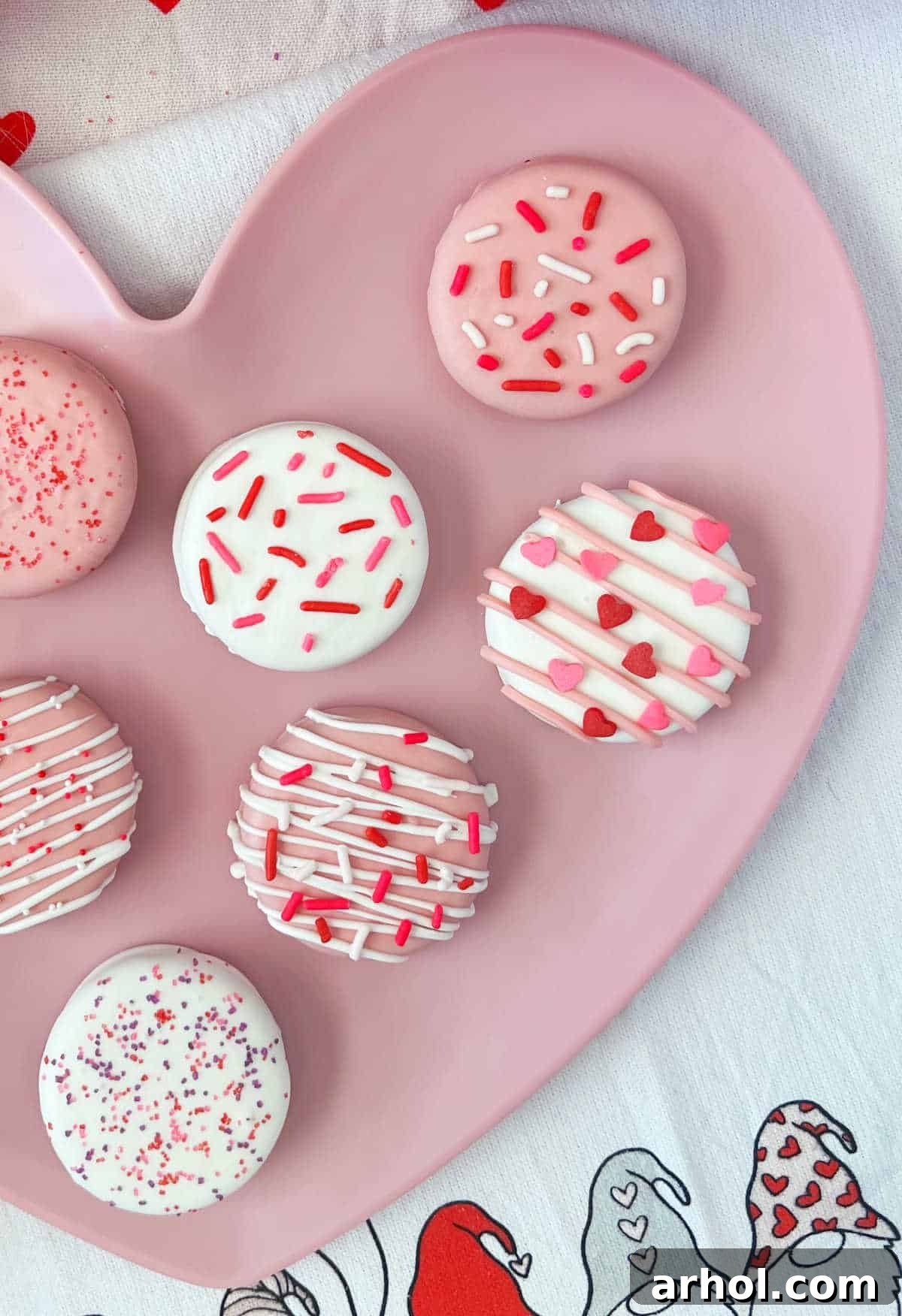 Irresistible White Chocolate Covered Oreos 6 A festive display of white chocolate dipped Oreos decorated with Valentine's Day sprinkles, resting on a charming gnome towel.