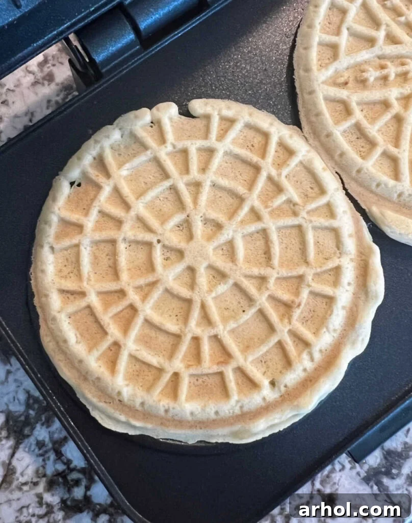 A freshly cooked pizzelle cookie being removed from the pizzelle iron, showing its golden-brown color and intricate pattern.
