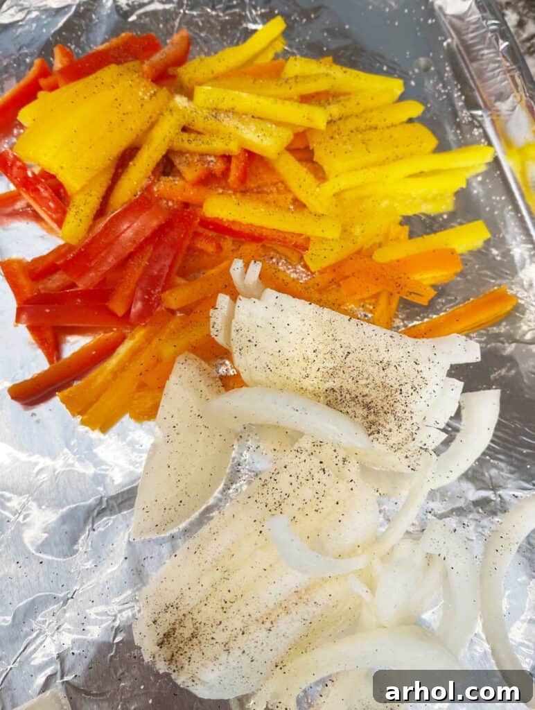 Thinly sliced bell peppers and red onion, seasoned with salt and pepper, spread evenly on a foil-lined baking sheet.