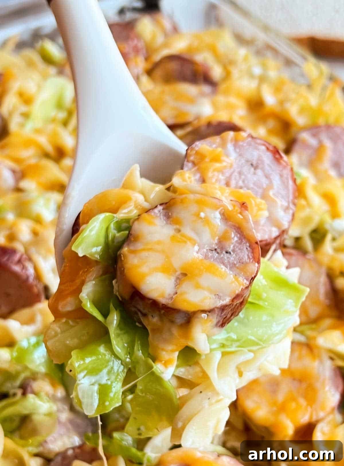 A serving spoon lifting cheesy smoked sausage, green cabbage, and pasta from a baking dish.
