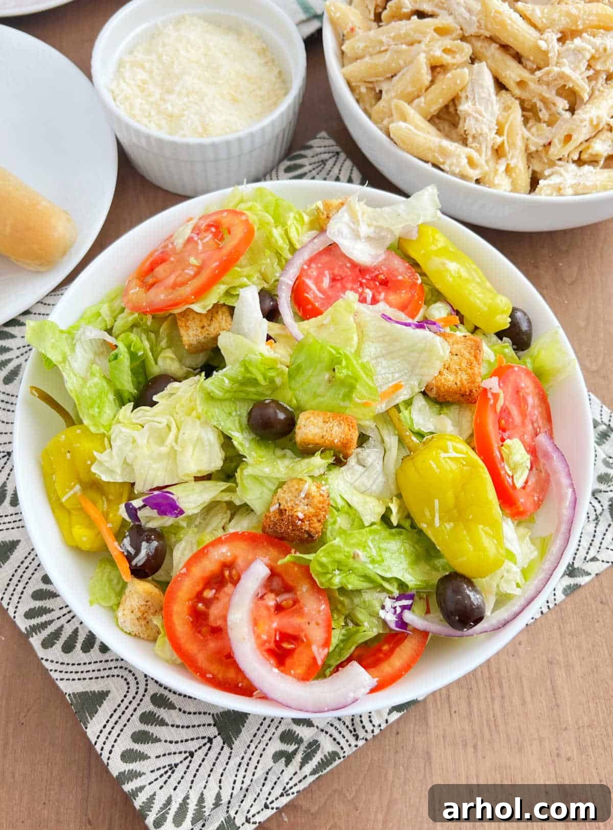 A large bowl of vibrant copycat Olive Garden salad, featuring crisp lettuce, sliced tomatoes, olives, red onion, and pepperoncinis, served on a rustic wooden table alongside a plate of pasta and golden breadsticks, evoking a cozy Italian dining experience at home.