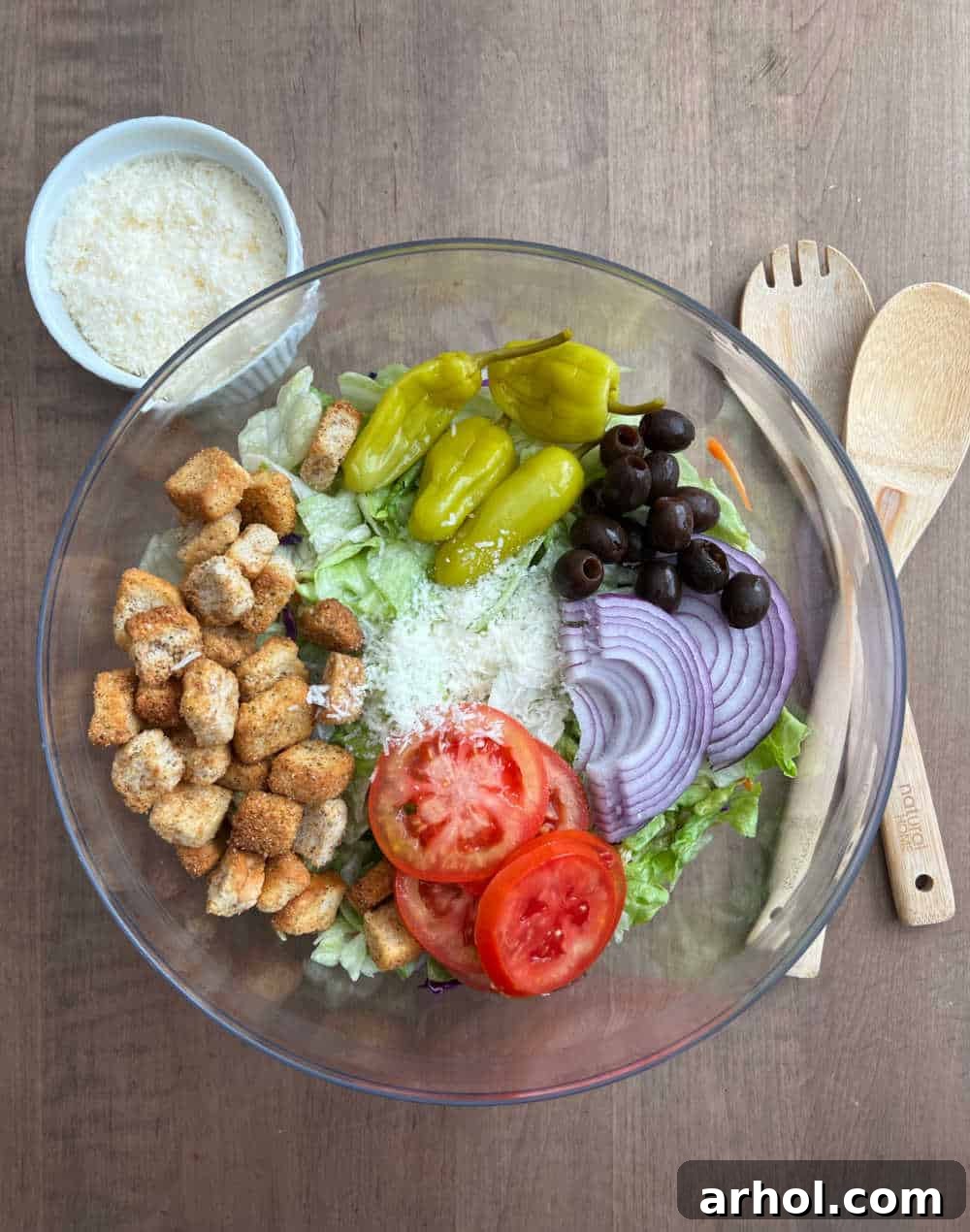 A large clear glass bowl filled with the base of a copycat Olive Garden salad, showing layers of crisp iceberg lettuce, bright red Roma tomato slices, black olives, thinly sliced red onion, and whole pepperoncini peppers, with a scattering of crunchy croutons and grated Parmesan cheese on top, ready for dressing.