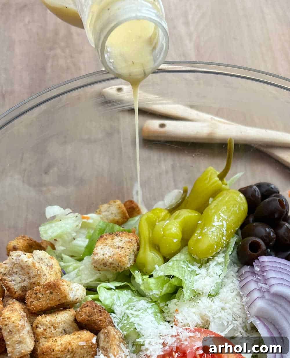 A close-up shot of an Olive Garden copycat salad in a glass bowl, with a hand pouring Olive Garden Italian dressing along the inner edge of the bowl, illustrating the technique for even dressing distribution.