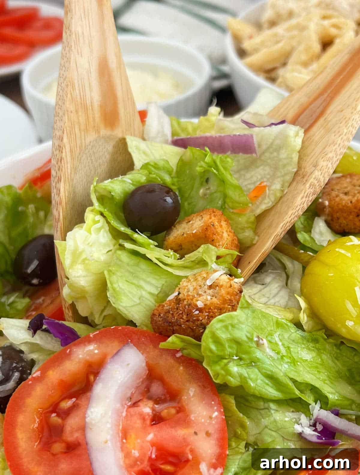 A pair of wooden salad serving spoons tossing a vibrant Olive Garden copycat salad in a large bowl, showcasing the fresh mixture of lettuce, tomatoes, olives, red onion, pepperoncinis, and croutons, all lightly coated in dressing, ready to be enjoyed.