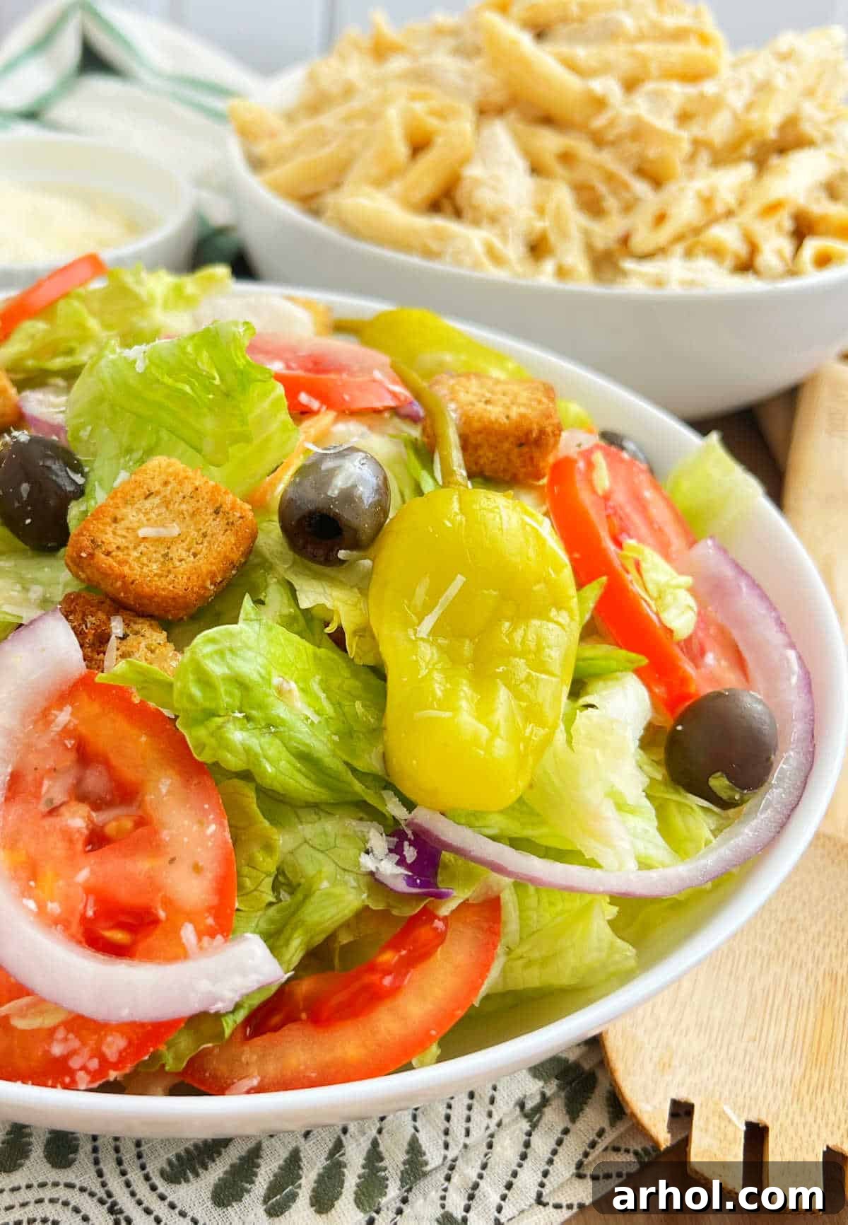 A generously sized serving bowl of copycat Olive Garden salad, artfully arranged to mimic the restaurant's presentation, featuring abundant crisp greens, colorful tomatoes, sliced red onion, black olives, and whole pepperoncinis, all glistening with dressing, served alongside a comforting pasta dish.