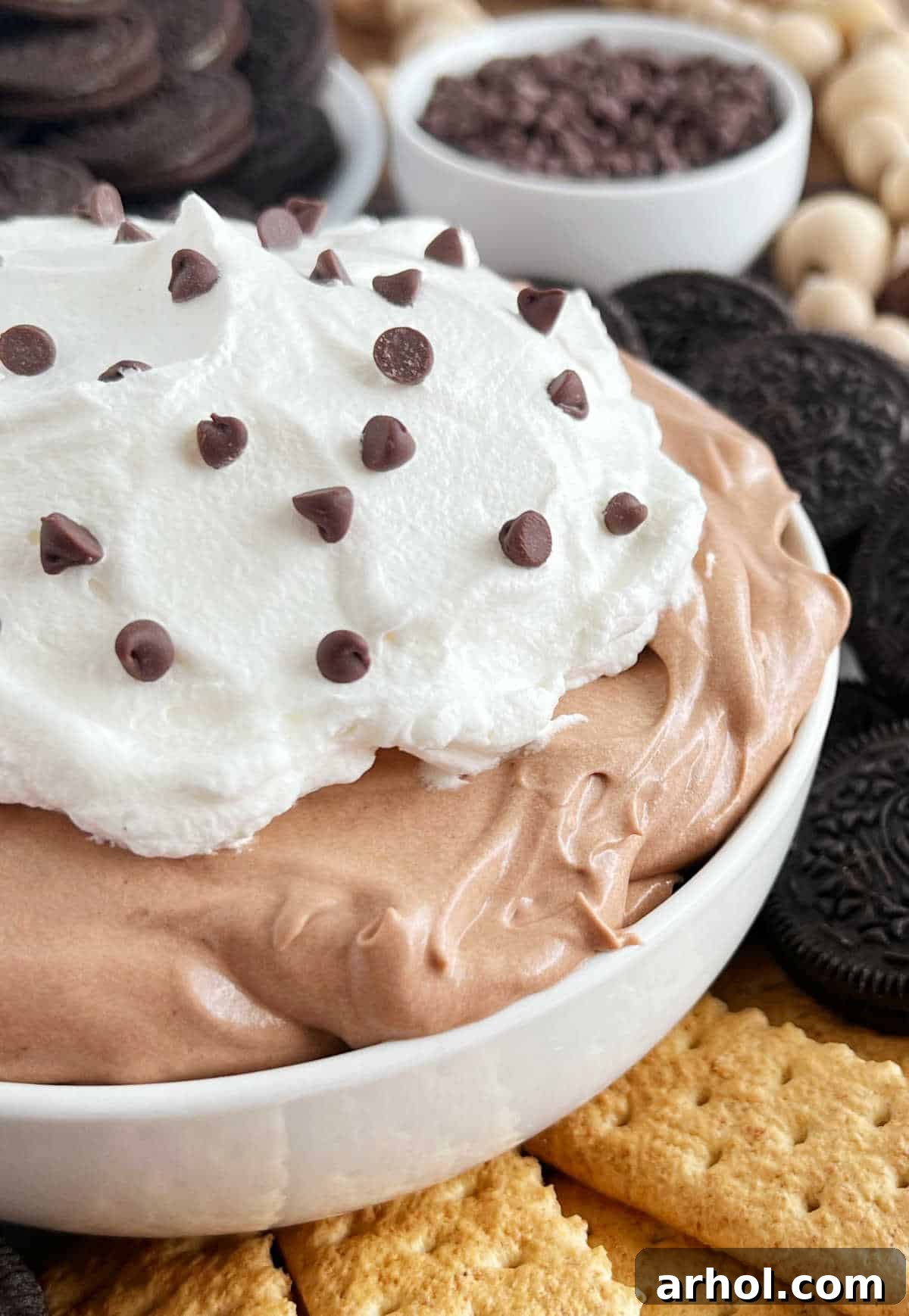 A bowl of creamy chocolate pudding pie dip, topped with whipped cream and chocolate chips, surrounded by various dippers like cookies and fruit.