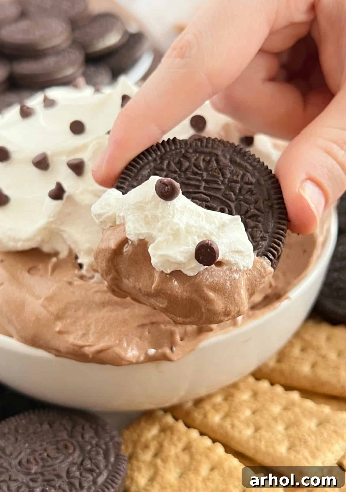 An Oreo cookie being dipped into a bowl of smooth chocolate pie dip, with other dippers in the background.