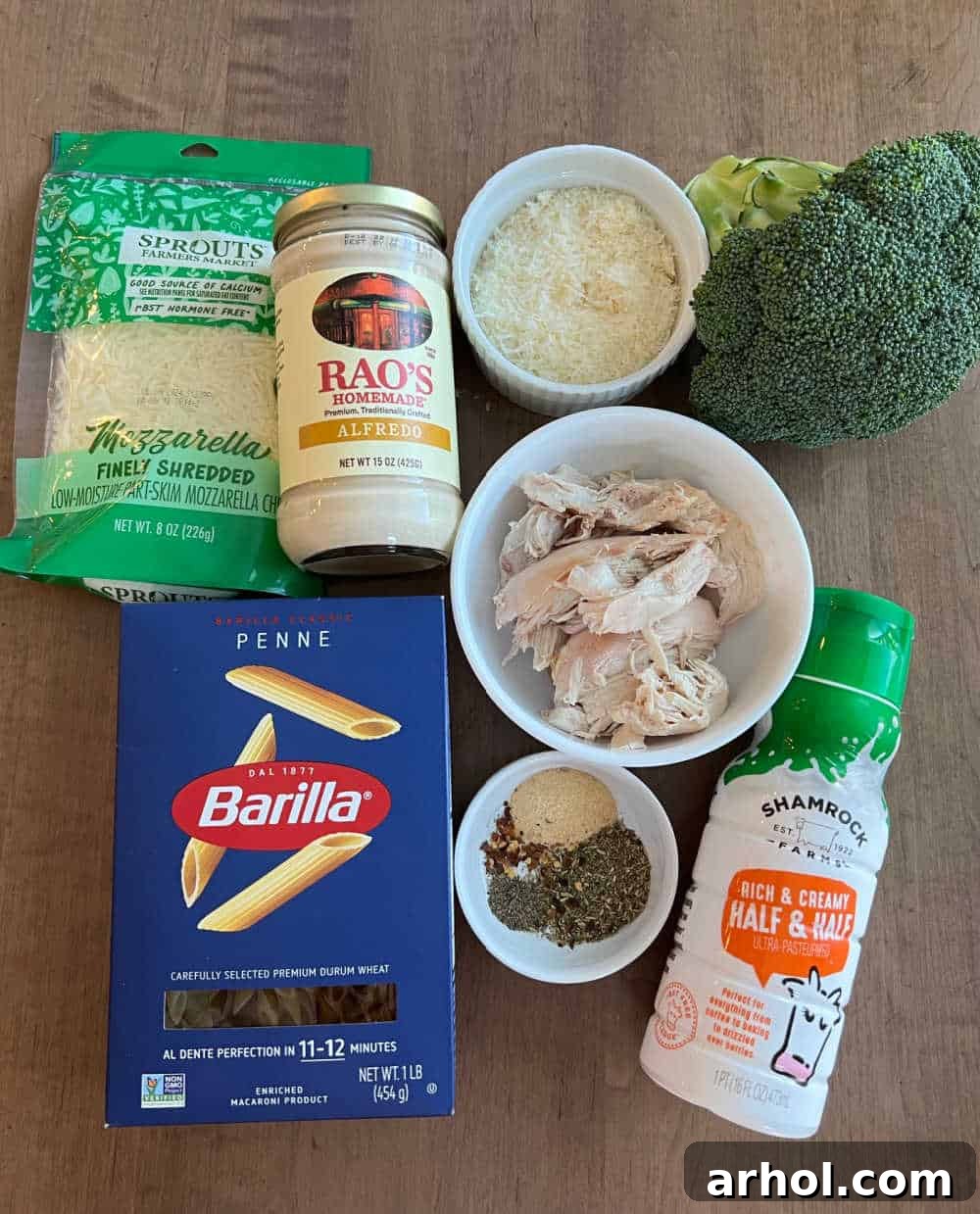 Key ingredients for Chicken Broccoli Alfredo Casserole including rotisserie chicken, penne pasta, Alfredo sauce, cream, fresh broccoli, assorted cheeses, and aromatic seasonings laid out on a kitchen counter.