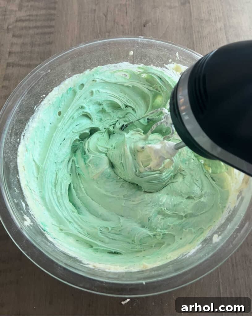 The process of mixing green cake mix cookie dough with an electric mixer in a spacious mixing bowl, ensuring even color and consistency.