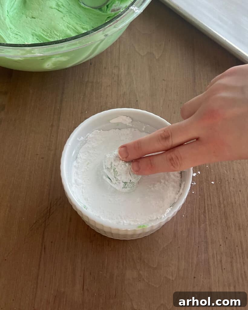 A hand carefully rolling a ball of green cookie dough generously in powdered sugar, preparing it for baking and the signature crinkle pattern.