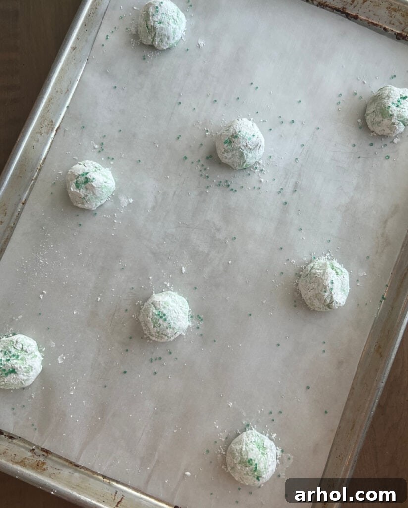 A baking sheet filled with evenly spaced green cookie dough balls, thoroughly coated in powdered sugar and ready for the oven.