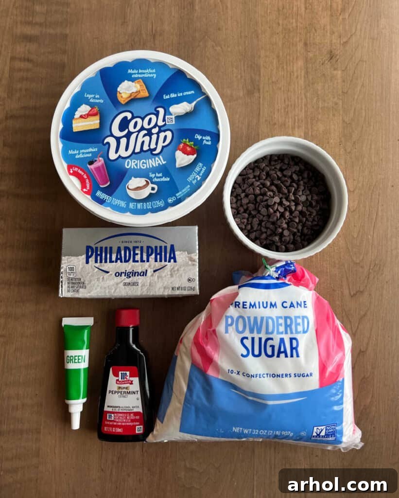 A flat lay of the key ingredients: thawed Cool Whip, softened cream cheese, green food coloring, peppermint extract, powdered sugar, and a bowl of mini chocolate chips.