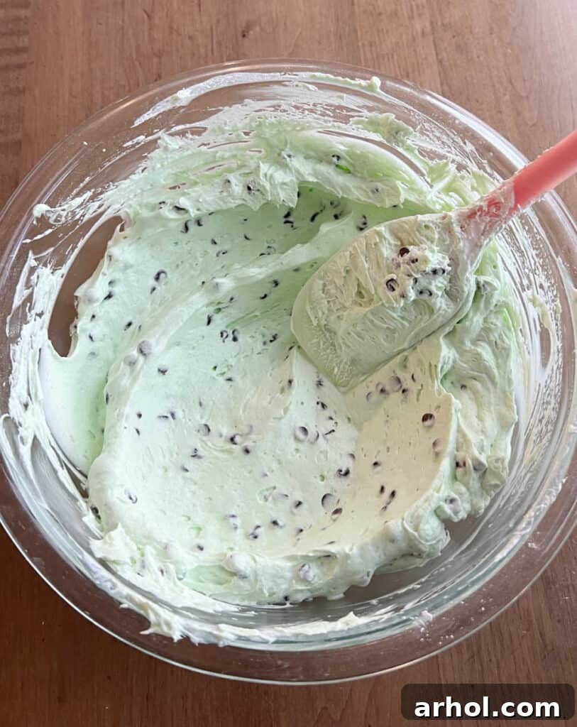 A close-up shot of mini chocolate chips being gently folded into the green mint cheesecake dip mixture with a rubber spatula.