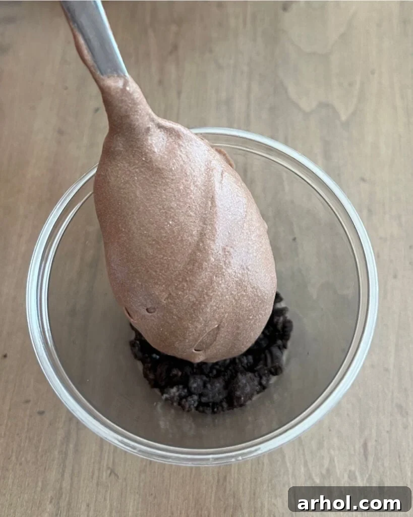 Spoonful of chocolate pudding mixture being added over a layer of crushed Oreo cookies in a clear cup.