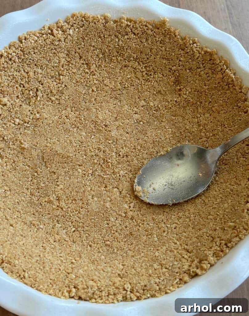 A close-up shot of crushed graham crackers being pressed firmly into a pie pan with the back of a spoon, creating the base for a delicious no-bake pie.