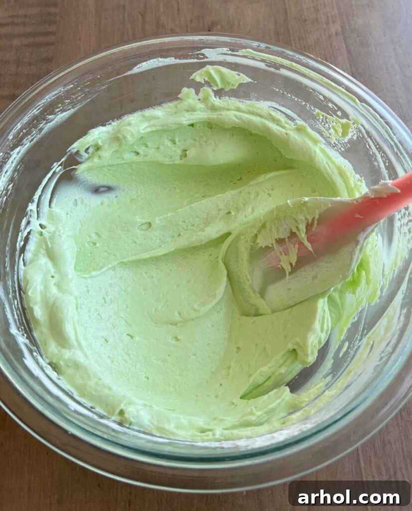 A rubber spatula gently folding thawed whipped topping into the green pudding mixture in a bowl, illustrating the technique to create a light and fluffy pie filling.
