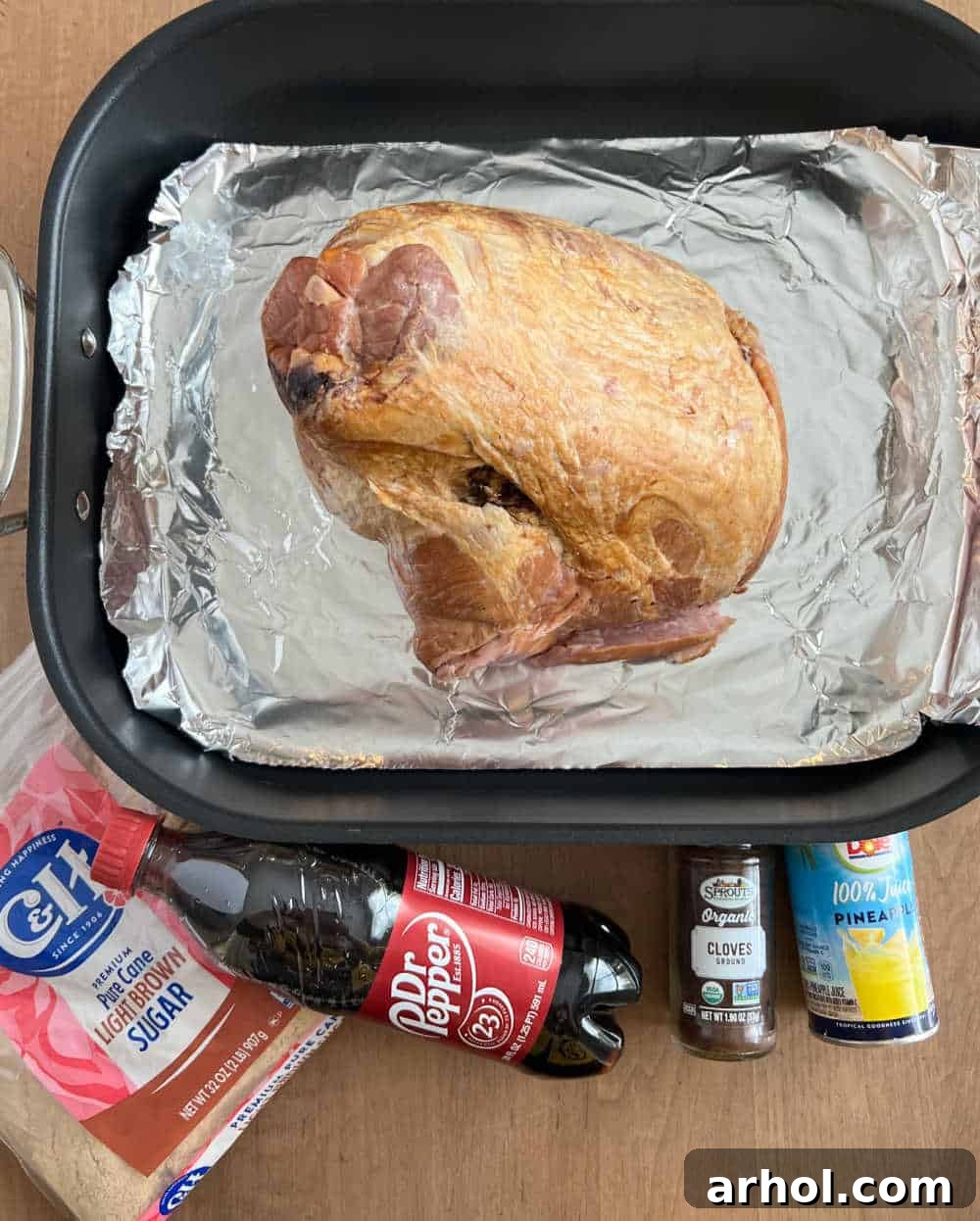 Uncooked spiral ham in a roasting pan, ready for the oven, surrounded by the glaze ingredients: brown sugar, Dr. Pepper, ground cloves, and pineapple juice.