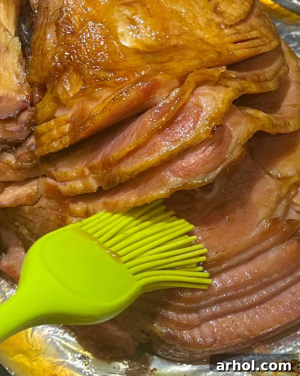 A hand brushing the glistening Dr. Pepper glaze over a baked spiral-cut ham in a roasting pan.