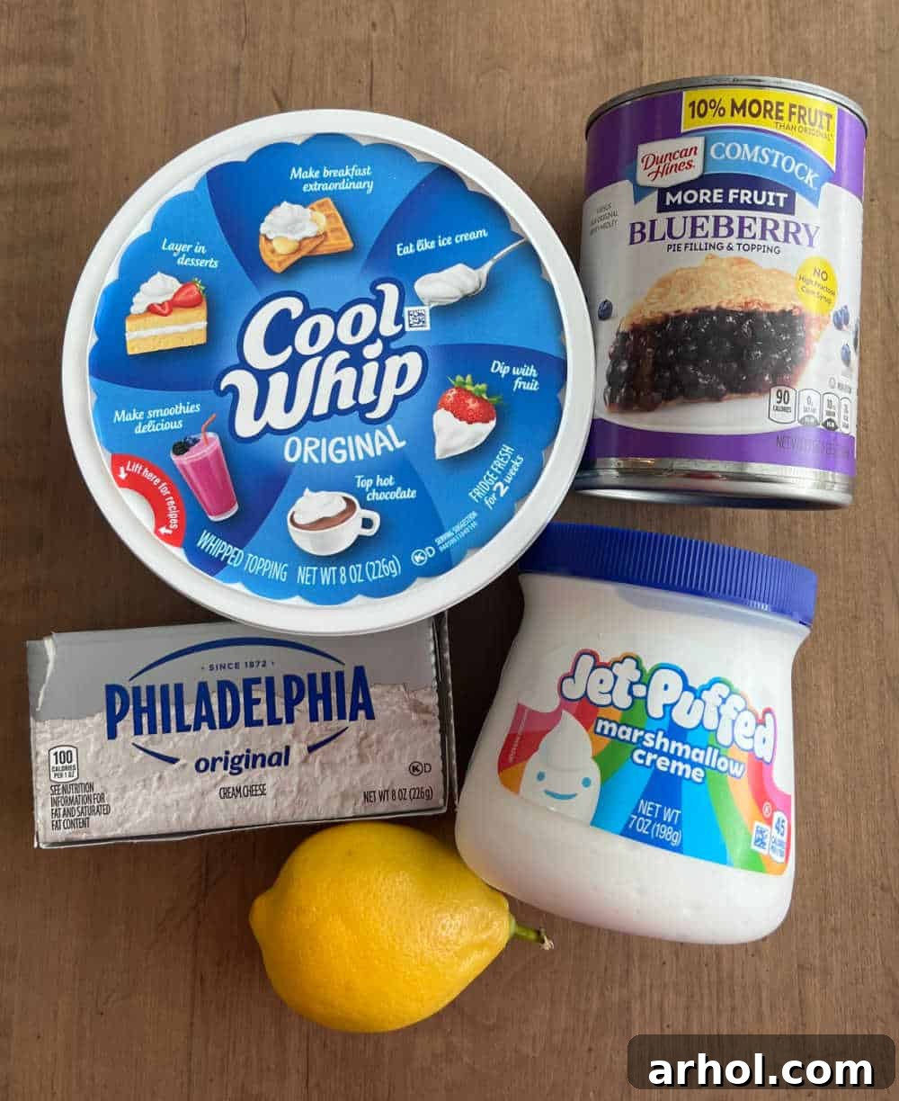 A flat lay of the key ingredients for lemon blueberry cheesecake dip: cool whip, blueberry pie filling, cream cheese, marshmallow fluff, and a fresh lemon, ready for preparation.