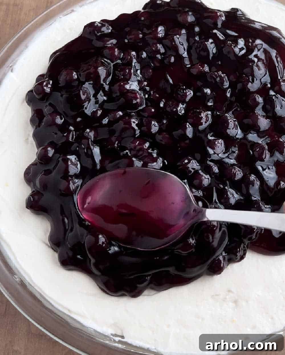 Spreading blueberry pie filling evenly over the creamy lemon cheesecake layer in a serving dish, ready for chilling and garnish.