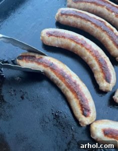 A large grilling spatula turning cooking sausage, peppers, and onions on the griddle.