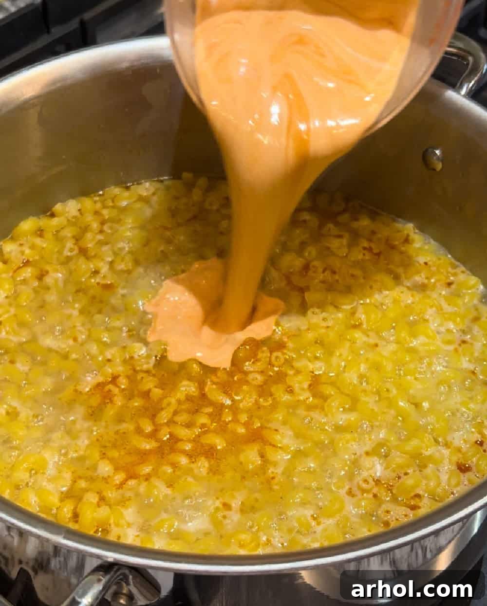 Pouring heavy cream and the remaining Buffalo sauce into macaroni noodles simmering in a pot.