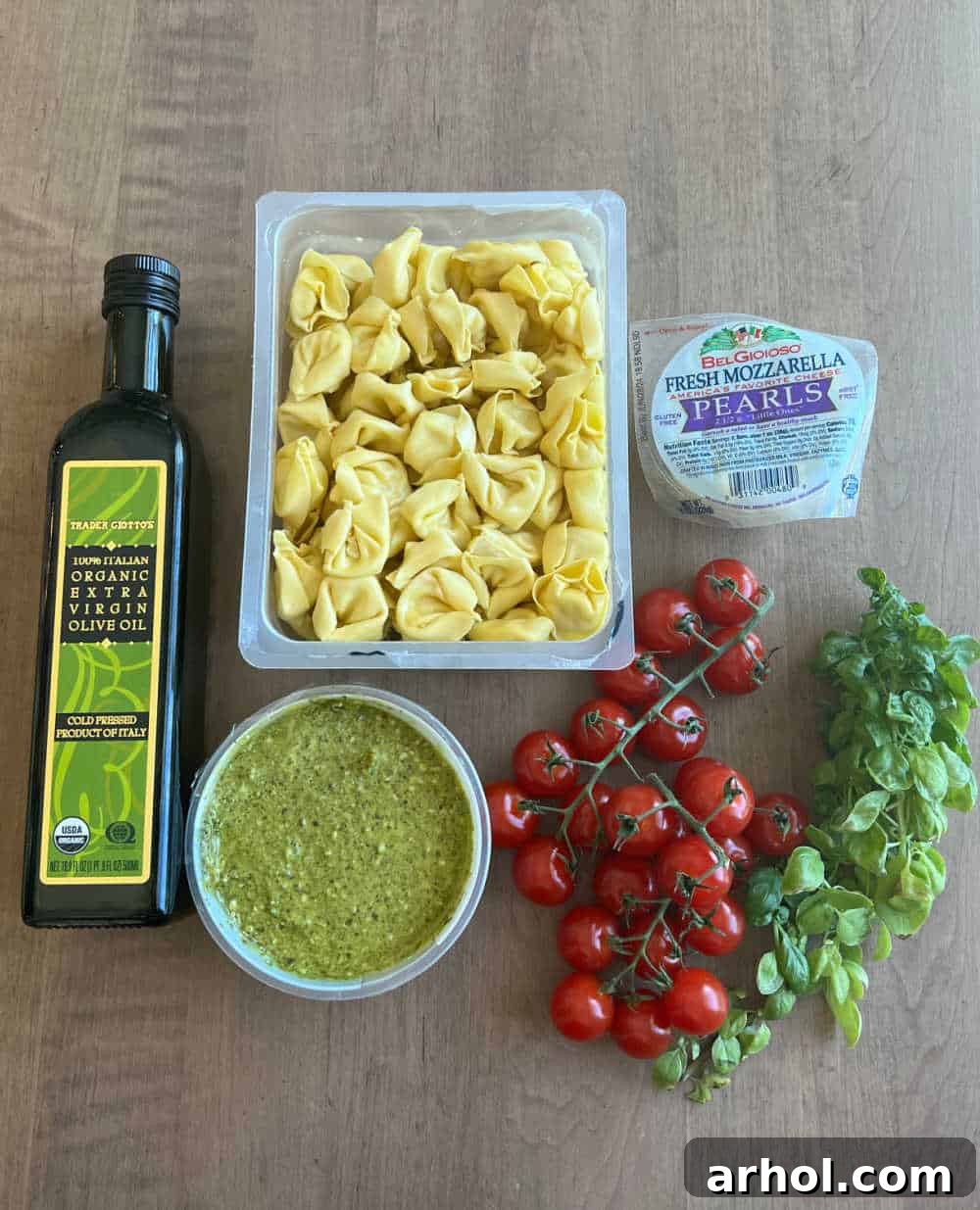 Close-up shot of the fresh ingredients for tortellini pasta salad: uncooked tortellini, cherry tomatoes, mozzarella pearls, basil pesto, fresh basil leaves, and a drizzle of olive oil.