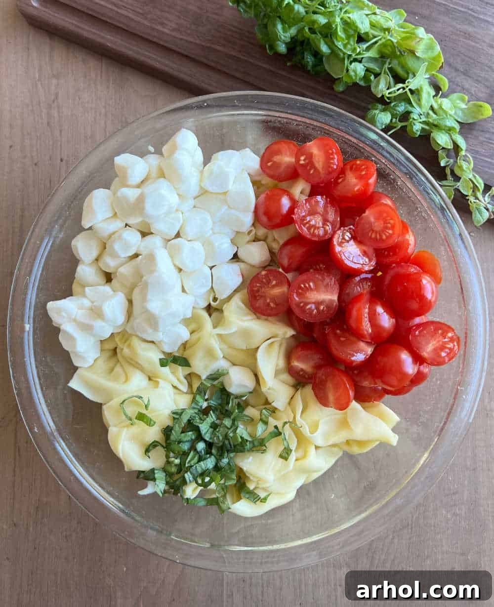 Freshly cooked tortellini, halved cherry tomatoes, mozzarella pearls, and chopped fresh basil leaves artfully arranged in a large mixing bowl, ready for the pesto.