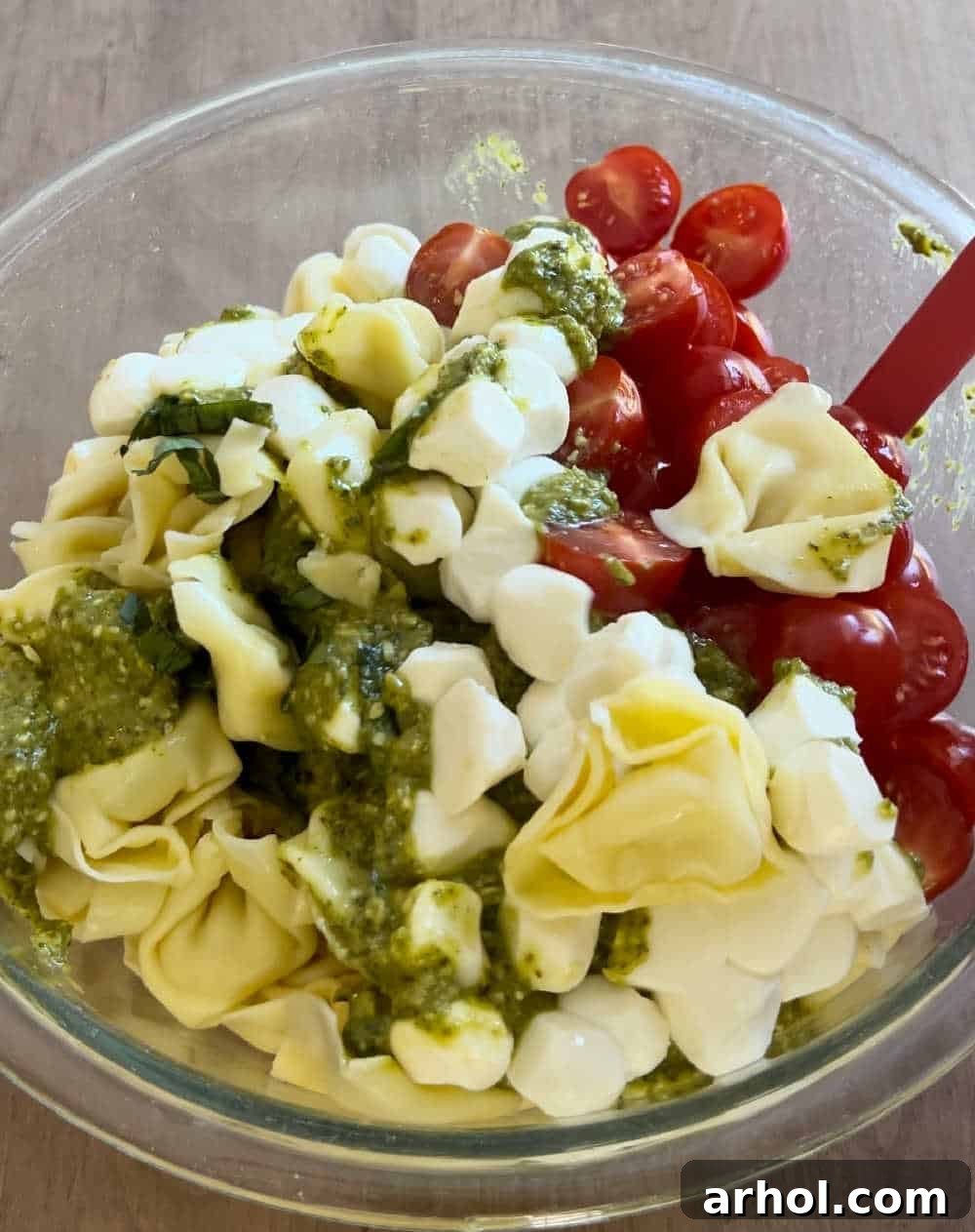 A large mixing bowl with tortellini pasta salad being gently stirred with a spoon, ensuring all ingredients are well-coated with the basil pesto sauce.