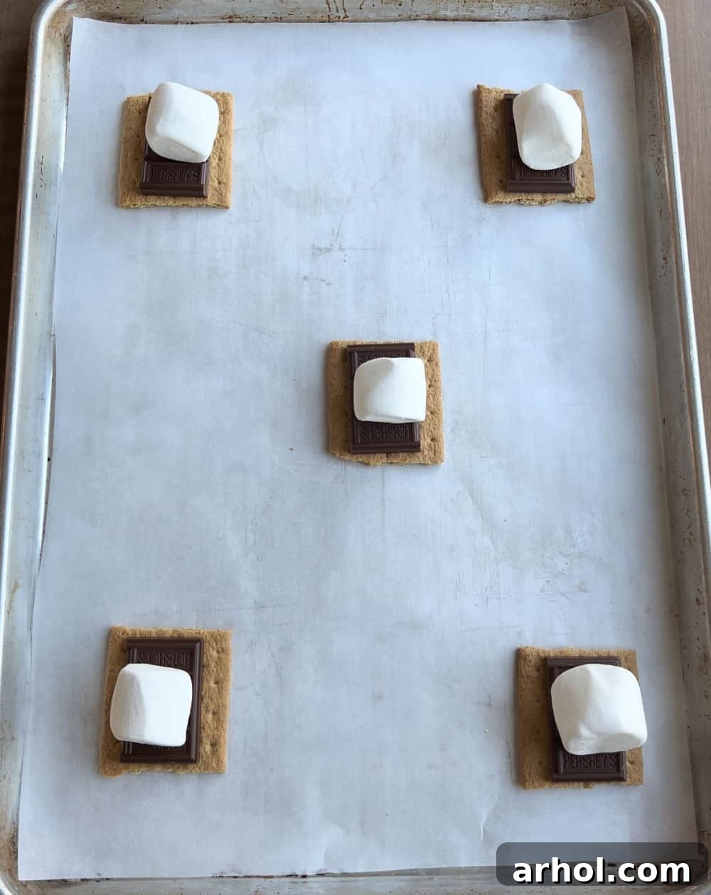Assembled S'mores cores on a parchment-lined baking sheet, featuring graham crackers topped with chocolate pieces and fluffy marshmallows.