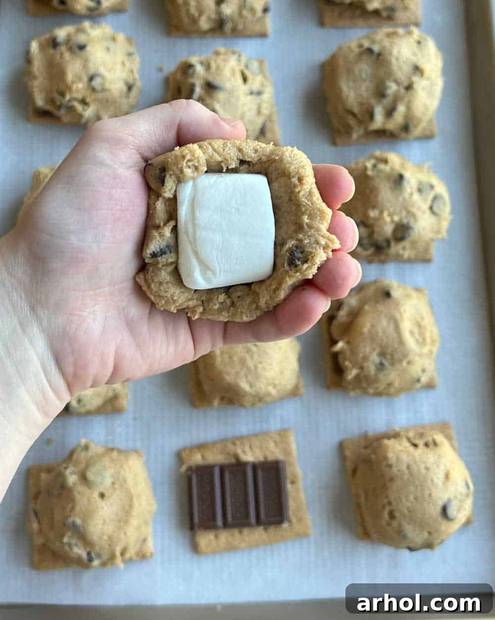 A close-up view of a large marshmallow being carefully wrapped and sealed with chocolate chip cookie dough, demonstrating the assembly process.