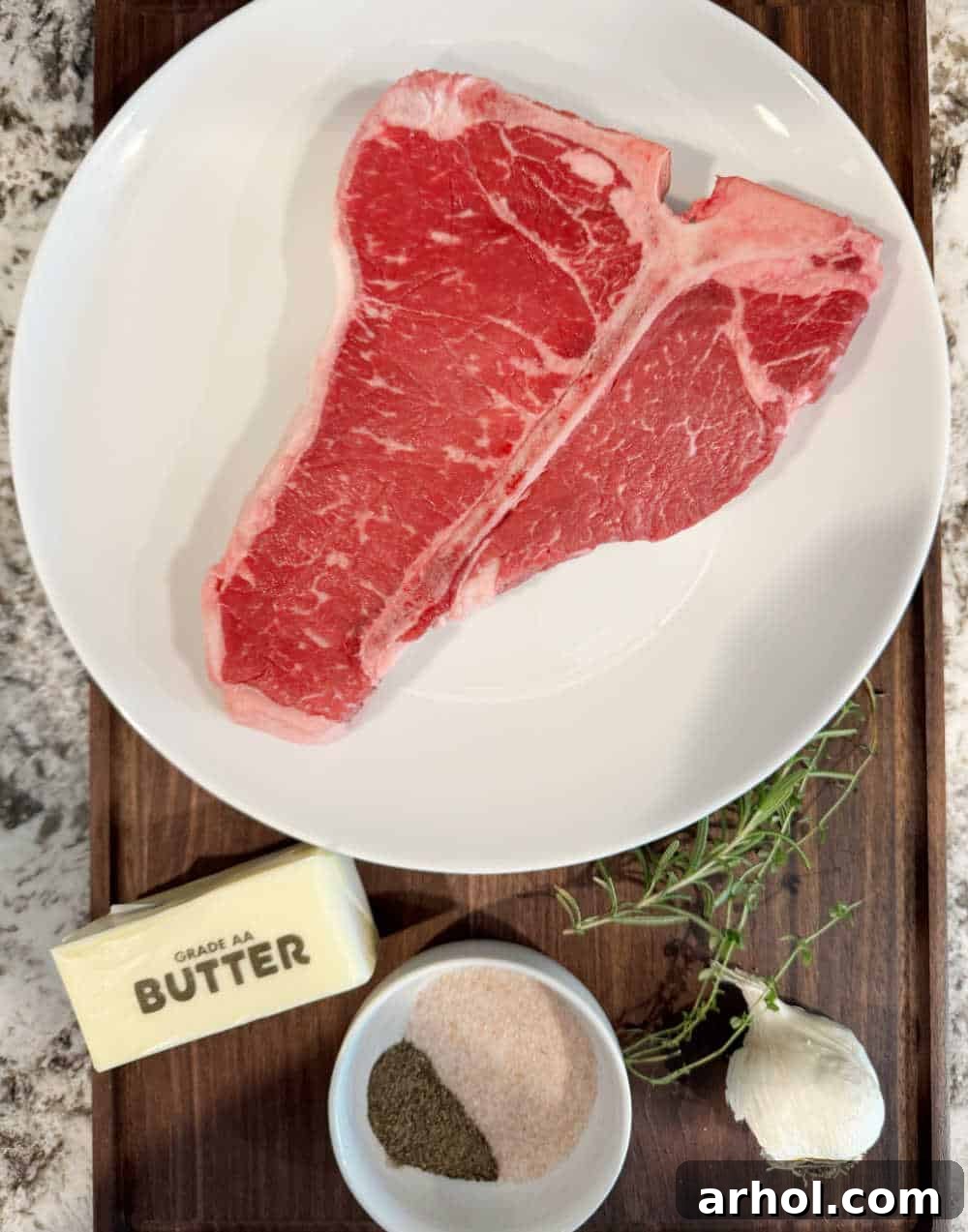 Raw porterhouse steak alongside unsalted butter, sea salt, black pepper, fresh garlic cloves, rosemary sprigs, and thyme sprigs on a rustic wooden cutting board.