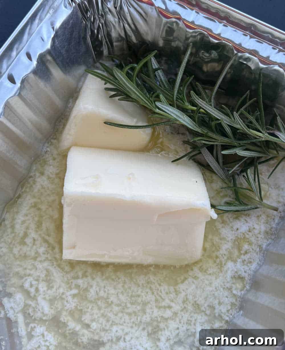 Melted butter, thinly sliced garlic, and a sprig of fresh rosemary simmering gently in a small aluminum foil pan on a low-heat zone of a Blackstone griddle.