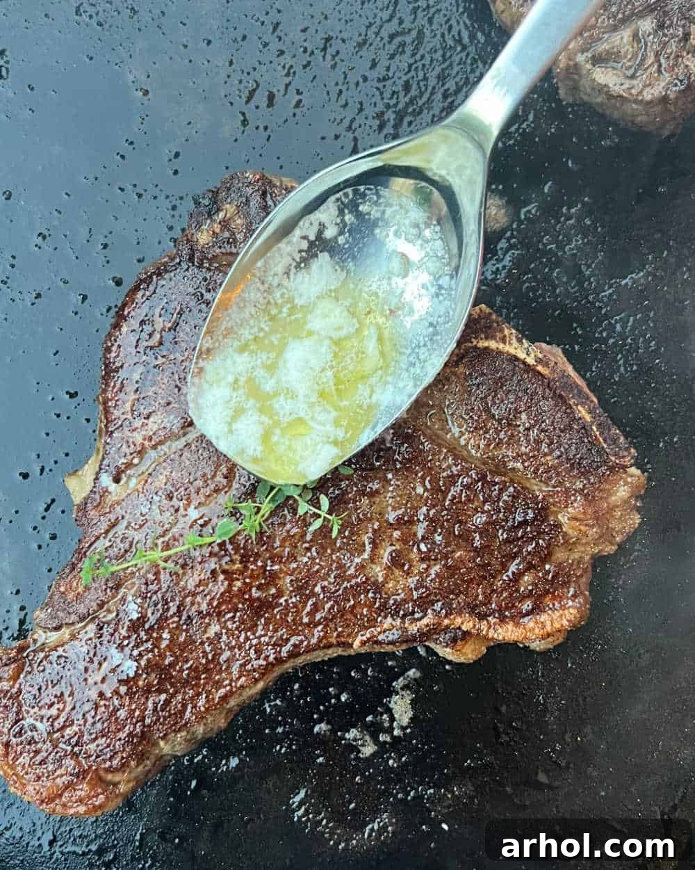 Grill tongs are used to baste sizzling steaks with melted garlic butter and herbs on a Blackstone griddle, enhancing flavor and moisture.