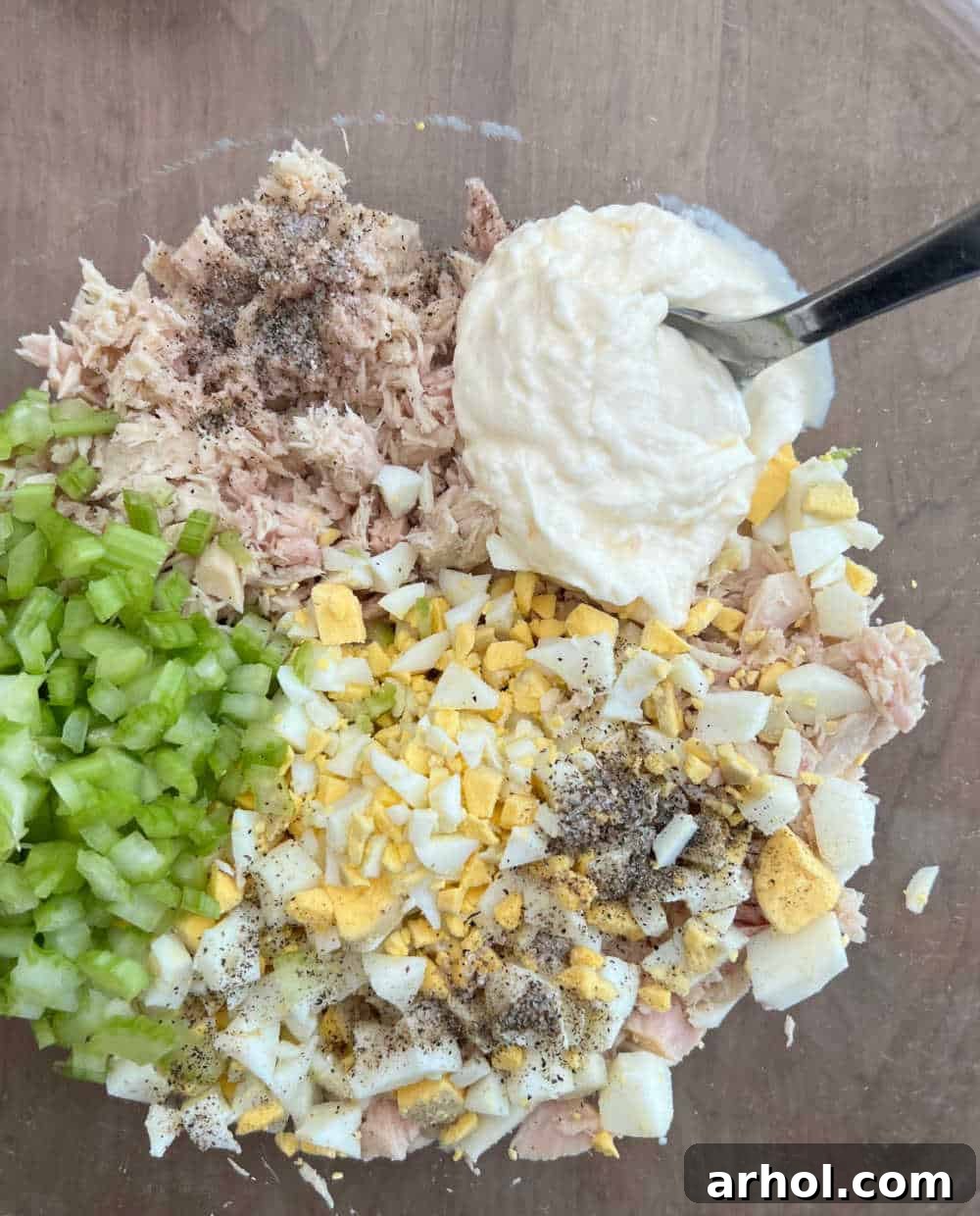 All the classic tuna salad ingredients, including tuna, chopped eggs, celery, and mayonnaise, being mixed together in a large bowl to create a harmonious blend.