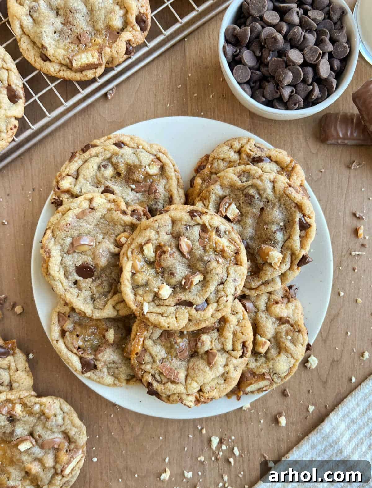 Plate of golden brown Twix chocolate chip cookies on a rustic wooden table, with chocolate chips and Twix pieces visible.