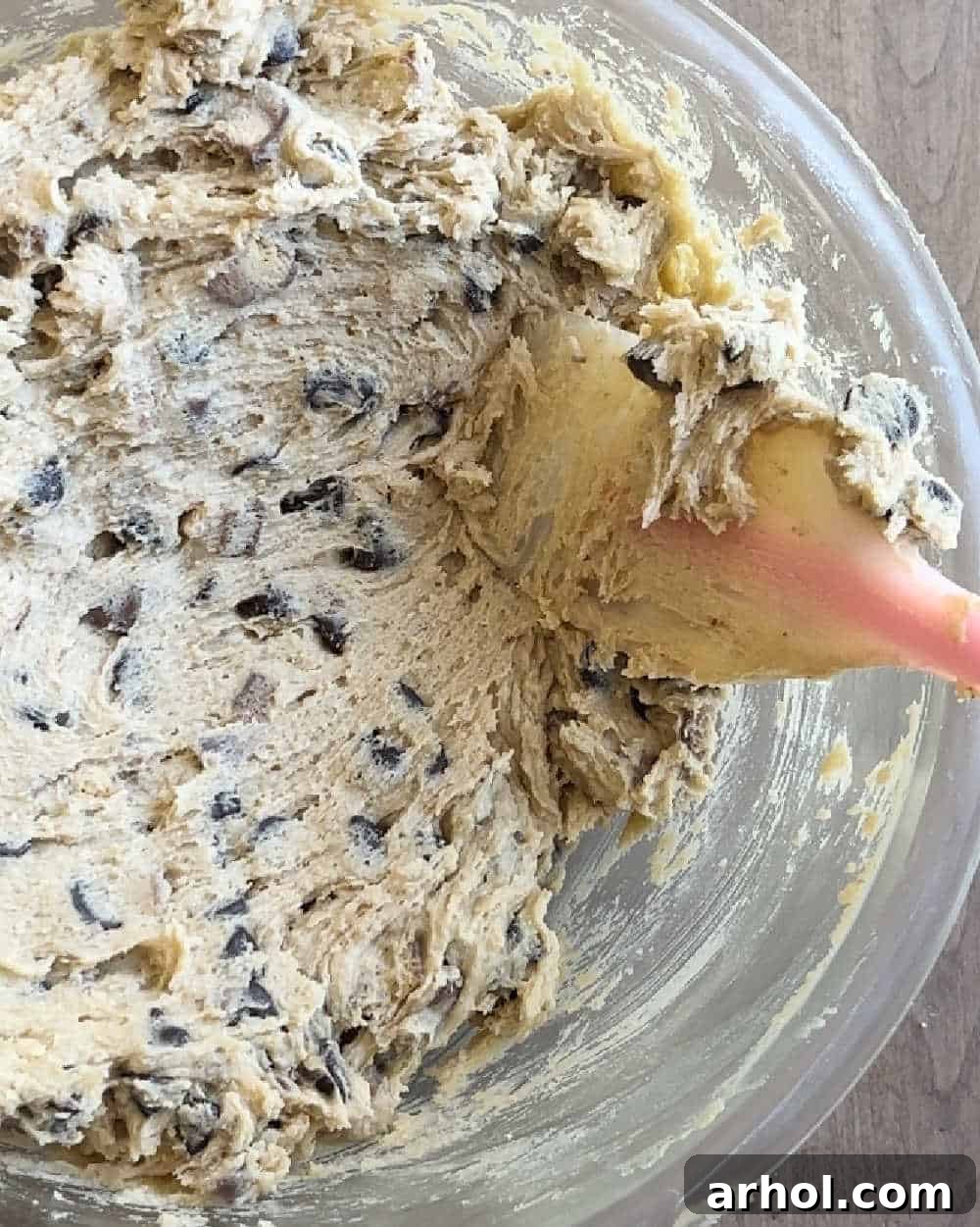 A hand stirring chocolate chips and chopped Twix candy bars into cookie dough with a spatula in a large mixing bowl.