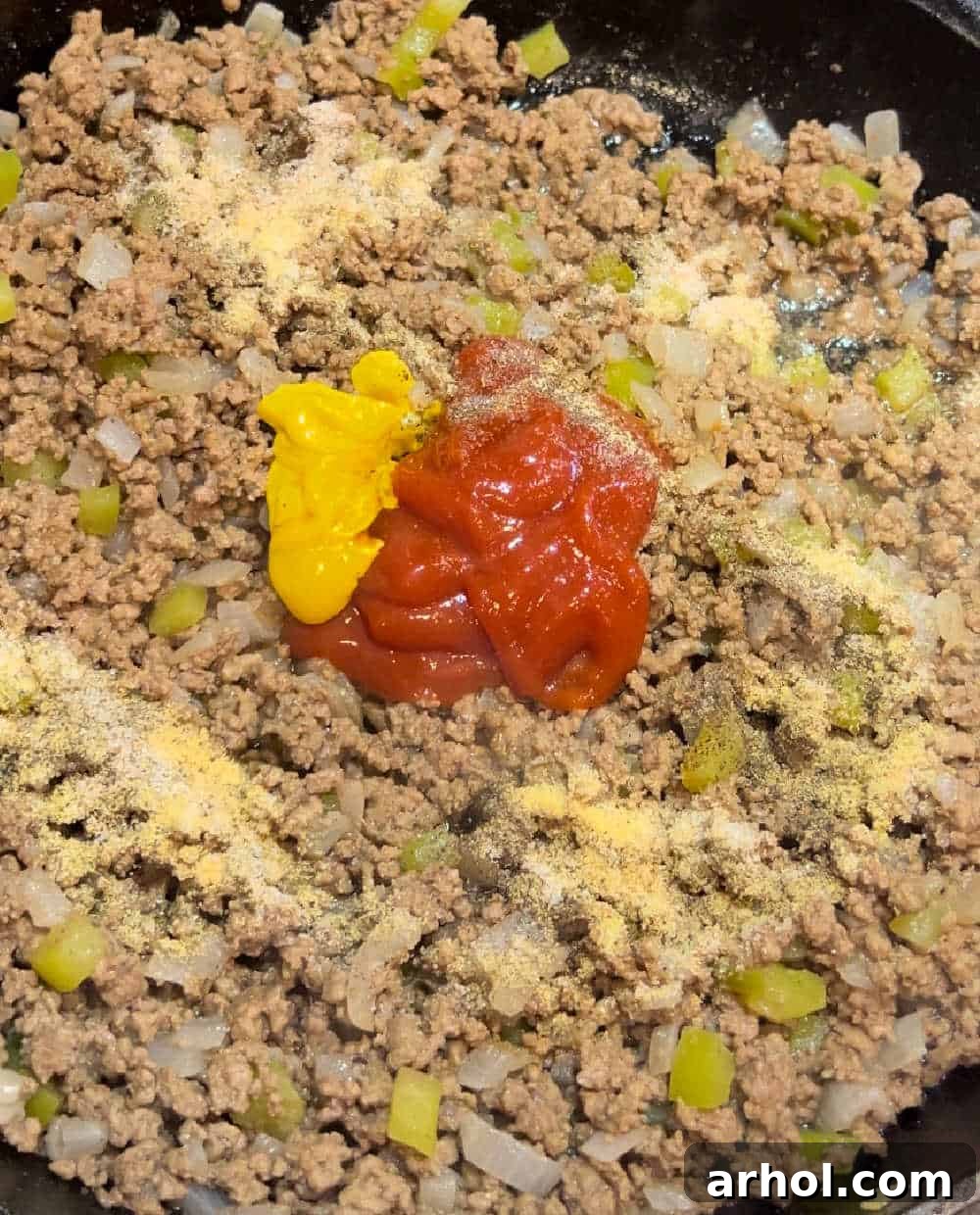 Adding ketchup, mustard, and seasonings to the cooked ground beef and vegetable mixture.