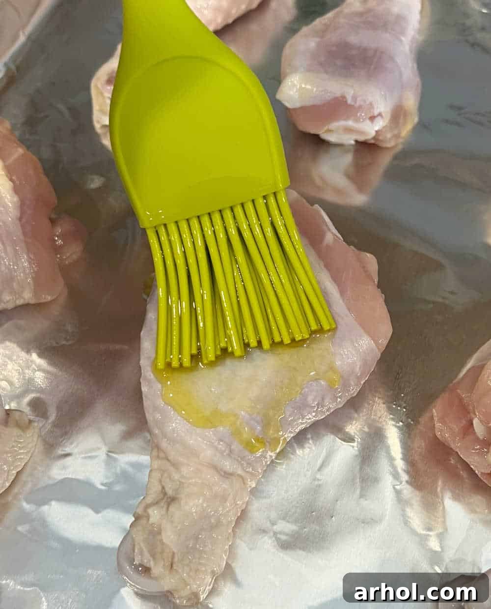 A close-up shot of raw chicken drumsticks arranged on a foil-lined baking sheet, being brushed with olive oil for preparation.