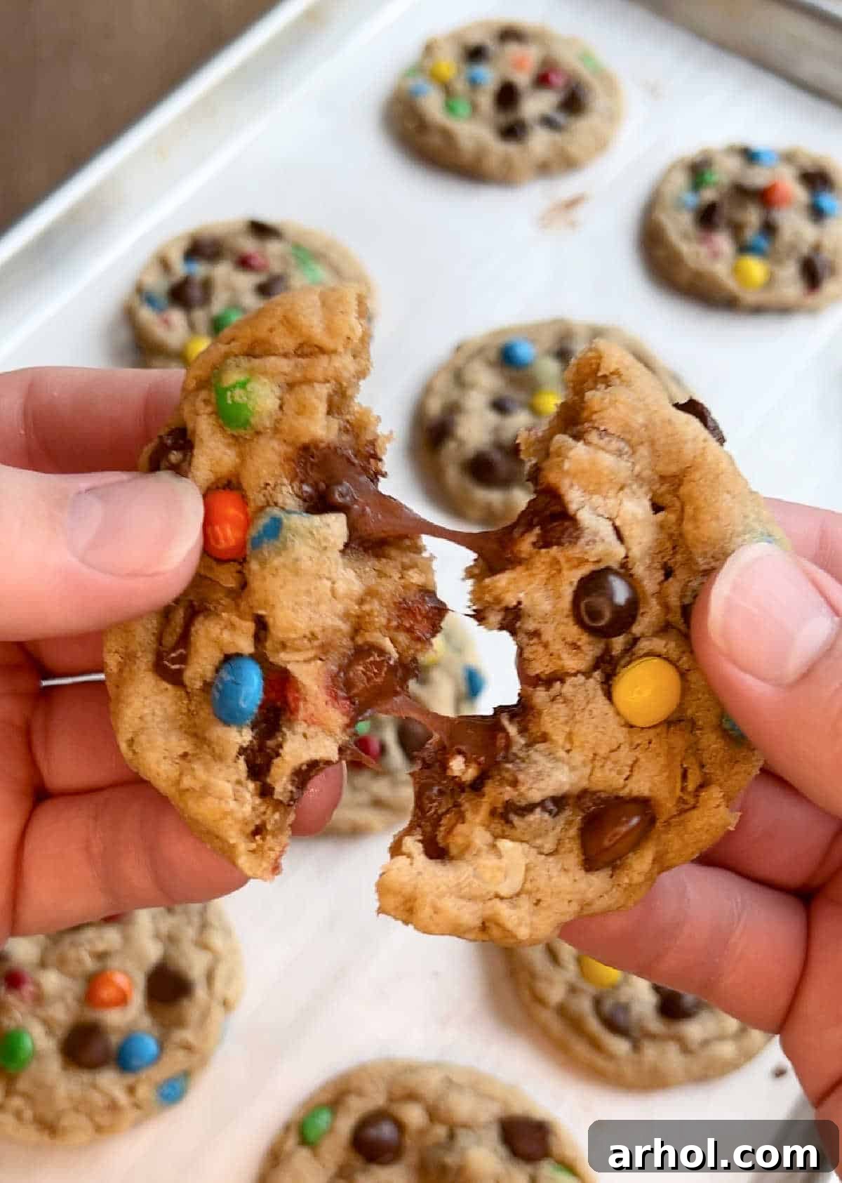 Hands pulling a warm, freshly baked monster cookie apart, showcasing its soft, chewy interior filled with mix-ins.