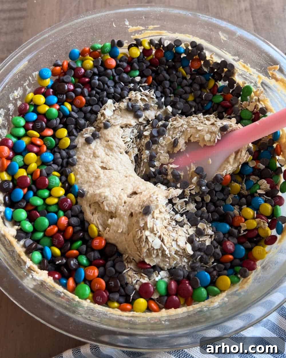 A hand mixing all the Monster Cookie Dough Dip ingredients together with a spatula in a large mixing bowl, showing the colorful candies and chocolate chips.
