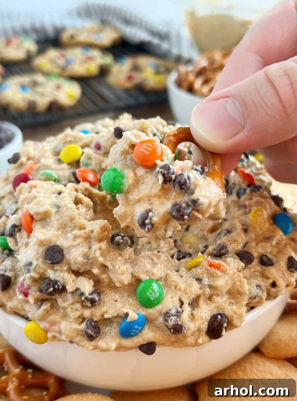 A close-up of a hand holding a mini pretzel generously loaded with Monster Cookie Dough Dip, showcasing the dip's rich texture and colorful mix-ins.
