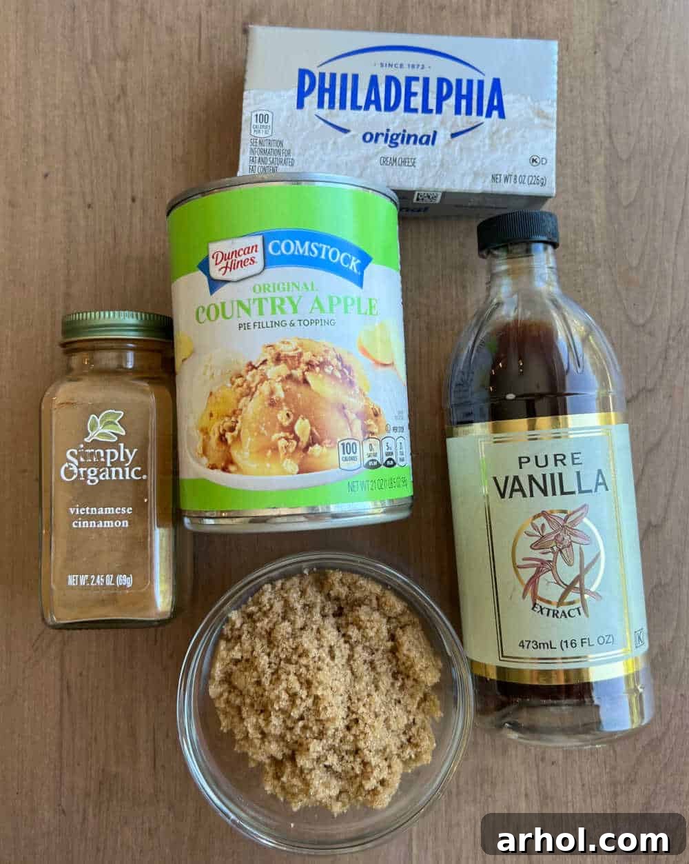 A pristine flat lay photograph showcasing the five simple, yet essential ingredients required for making a delicious apple pie dip: a block of softened cream cheese, rich brown sugar, fragrant vanilla extract, warming ground cinnamon, and a convenient can of apple pie filling.